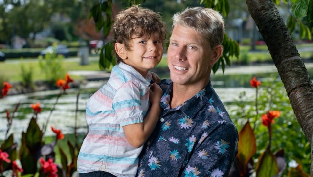Jason Bitzer holding his son James, a young neuroblastoma patient, by a pond surrounded by flowers — highlighting the human side of pediatric cancer and access to treatment.
