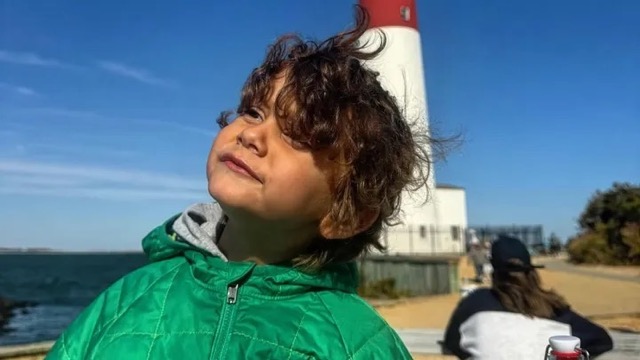 James Bitzer, a 5-year-old neuroblastoma patient, smiling in a green jacket near a lighthouse — symbolizing resilience and the fight for access to pediatric cancer treatment.